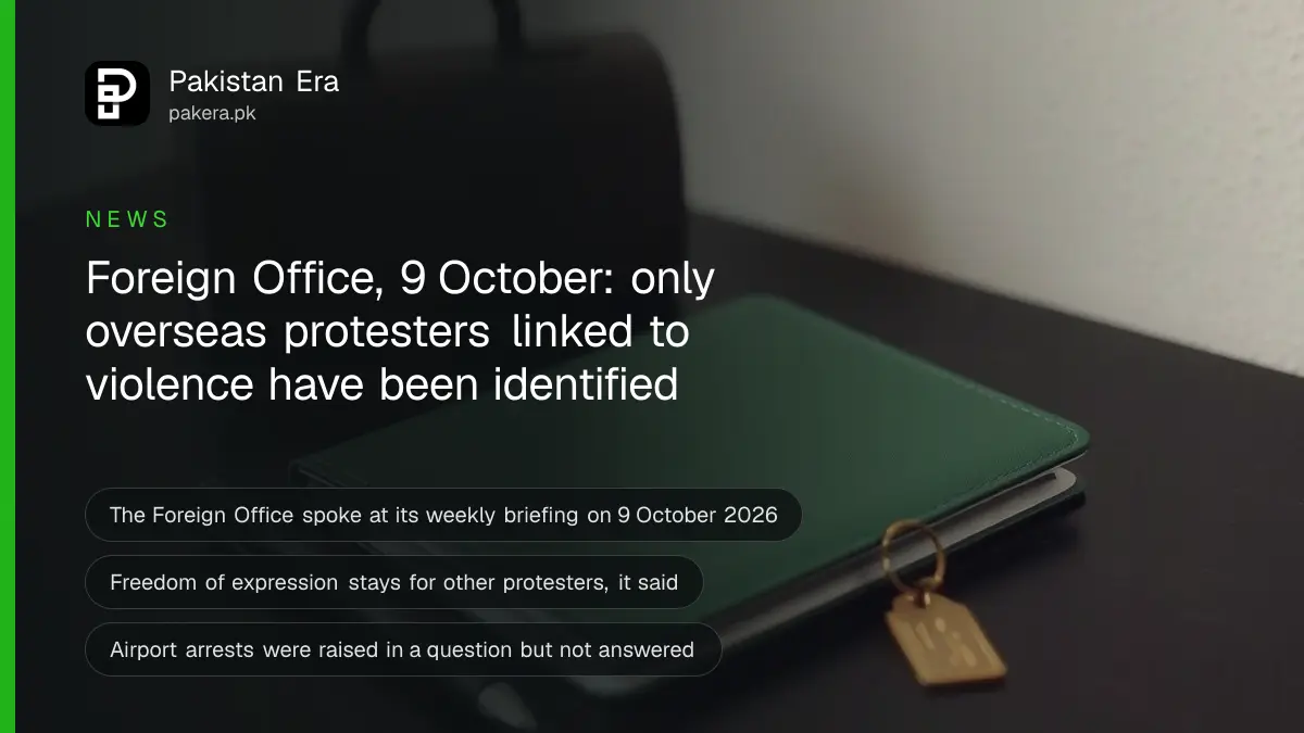 A closed green passport, a pen and a small brass luggage tag on a dark wooden table against a plain cream wall