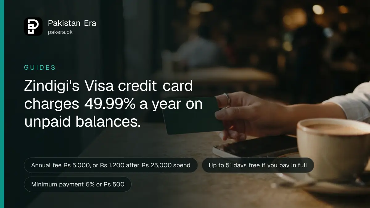 A hand holding a blank payment card beside a smartphone on a desk in Karachi